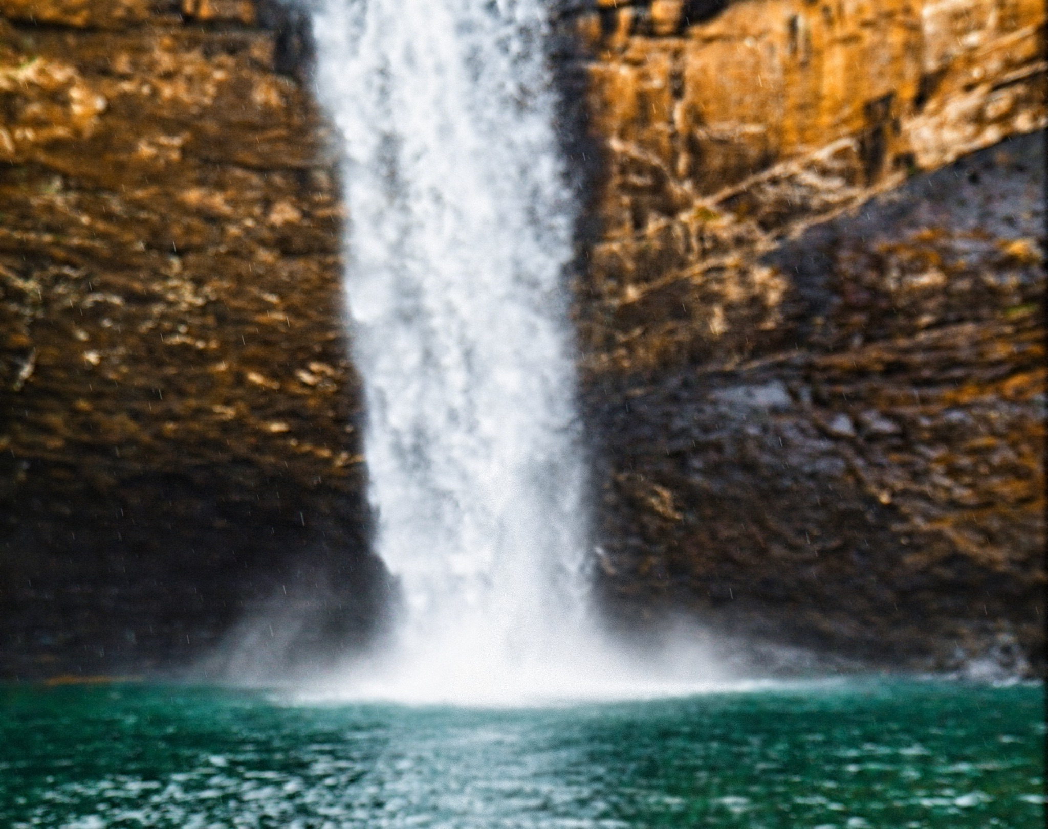 A tall waterfall pouring into a green pool between golden sandstone cliffs